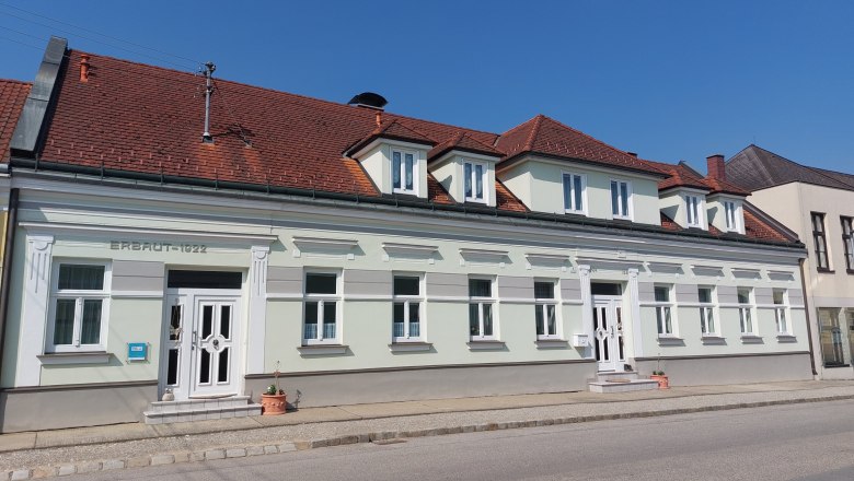 Ein zweistöckiges Gebäude mit rotem Ziegeldach und weißen Fensterrahmen, erbaut 1922, bei sonnigem Wetter.
