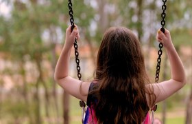 Ein Kind schaukelt im Freien, von hinten gesehen, umgeben von Bäumen.