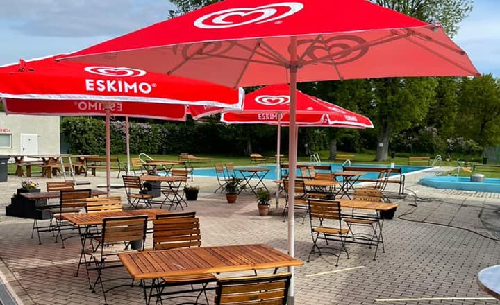 An outdoor pool with red parasols and empty wooden tables on a paved terrace.