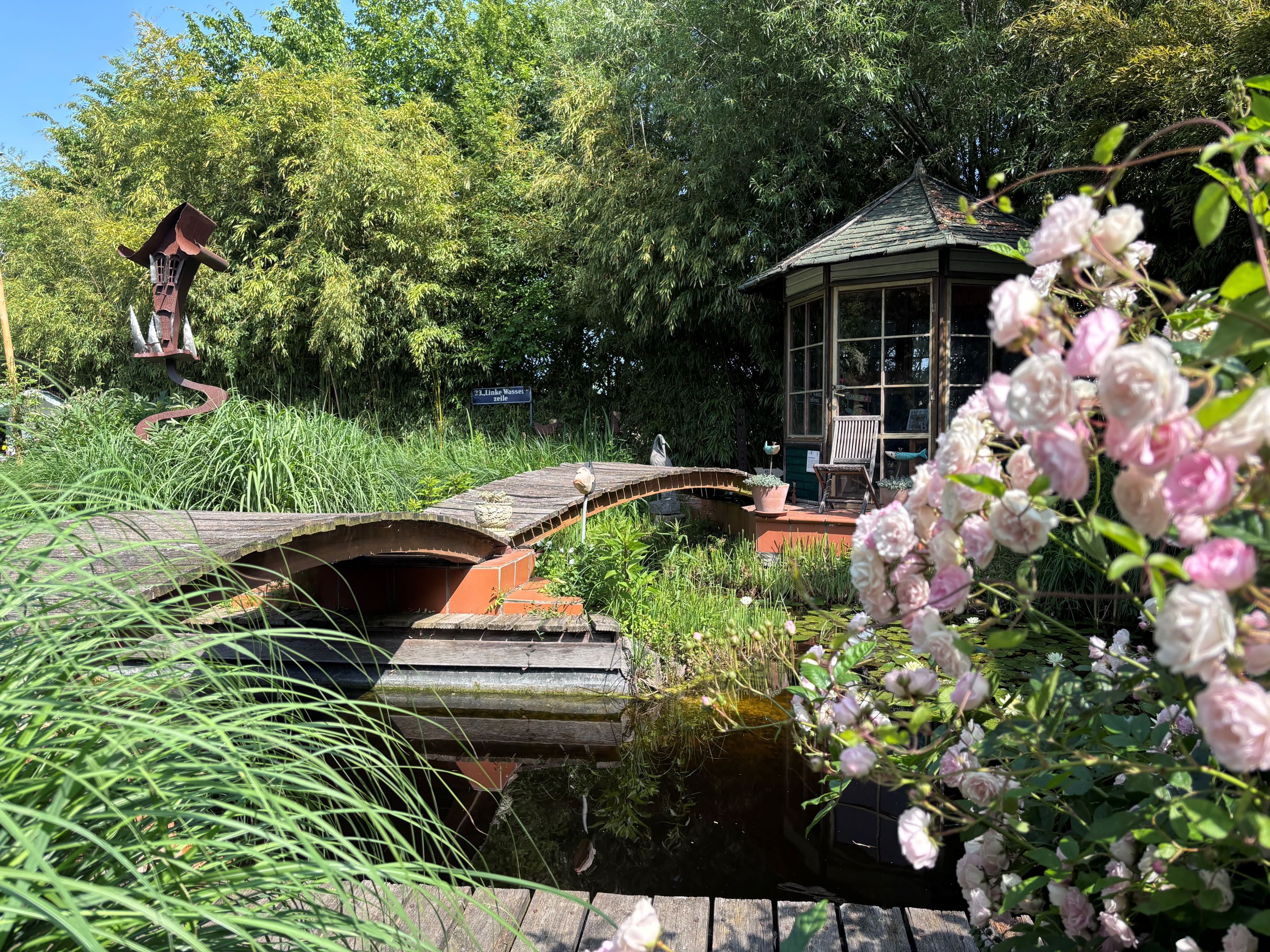 Ein idyllischer Garten mit Teich, Brücke, Pavillon und blühenden Rosen im Vordergrund.