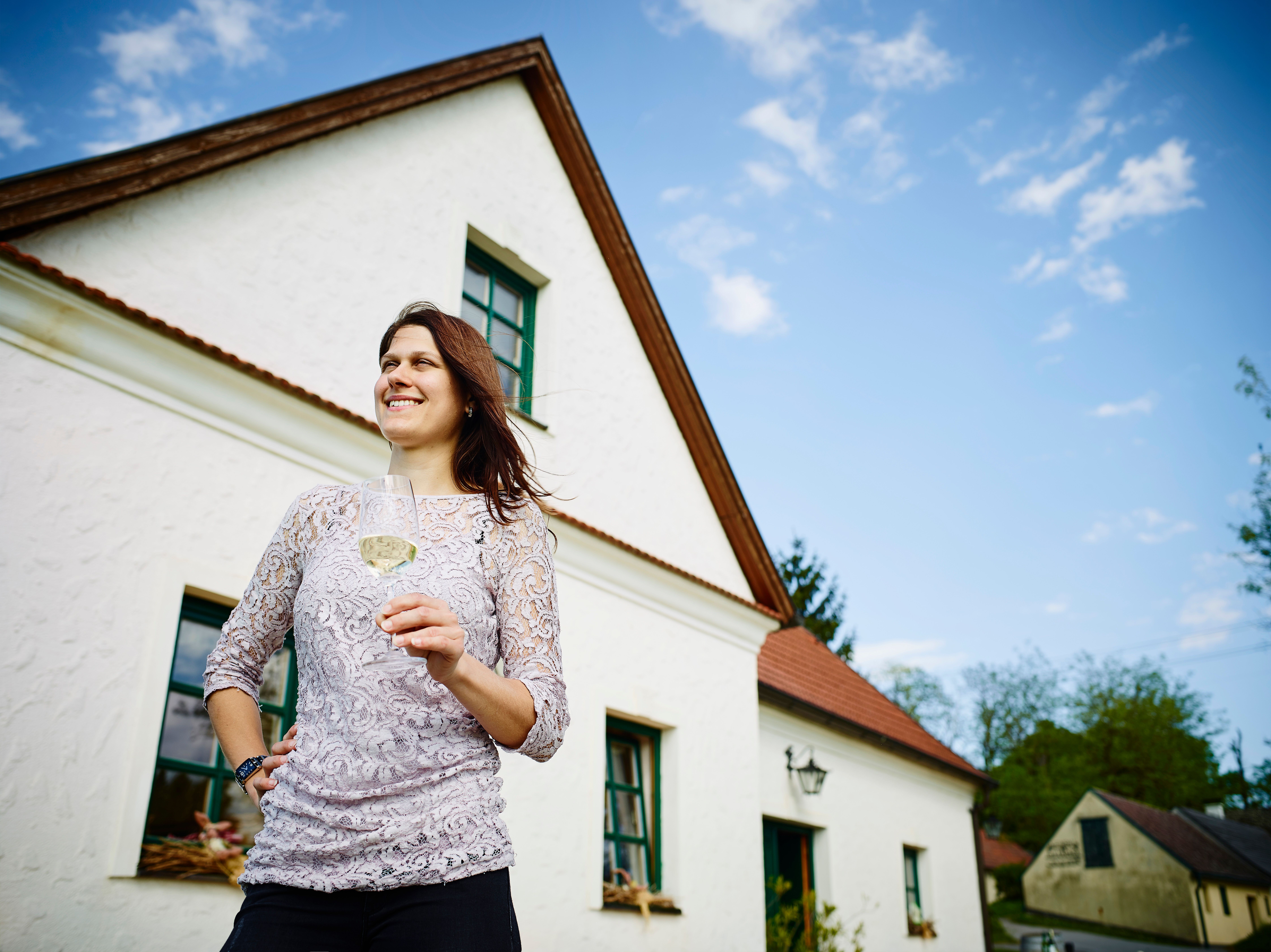 Frau mit Weinglas vor einem weißen Haus mit rotem Dach.