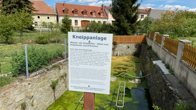 Kneippanlage mit Infotafel und Wasserbecken, umgeben von Zaun und Häusern im Hintergrund.