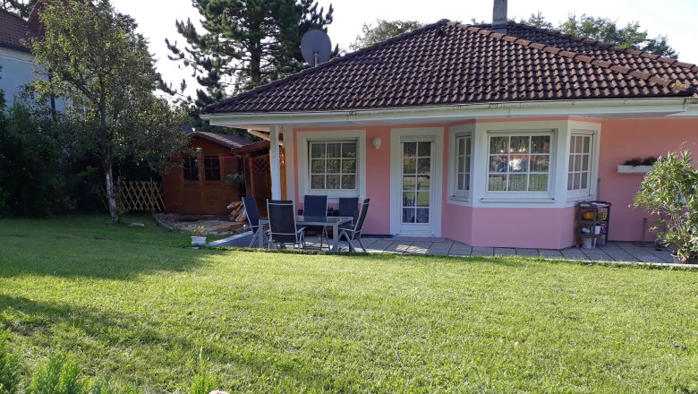 Haus Muck, © Barbara Muck Ein rosa Haus mit Garten, Terrasse und Gartenmöbeln, umgeben von Bäumen und Blumen.