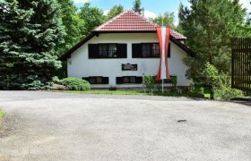 Ein weißes Haus mit rotem Dach und österreichischer Flagge davor, umgeben von Bäumen.