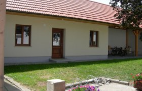 Ein Innenhof mit einem Gebäude mit rotem Ziegeldach, Holztür und Fenstern. Im Vordergrund sind Blumen und ein Fahrradständer zu sehen.