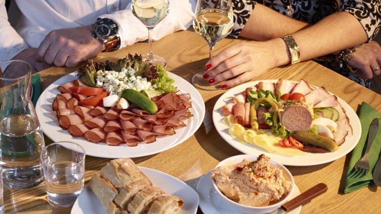Zwei Personen sitzen an einem Tisch mit Wurst- und Käseplatten, Brot und Wein.