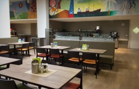 Innenansicht eines Restaurants mit leeren Tischen und Stühlen, bunte Wandmalerei im Hintergrund.