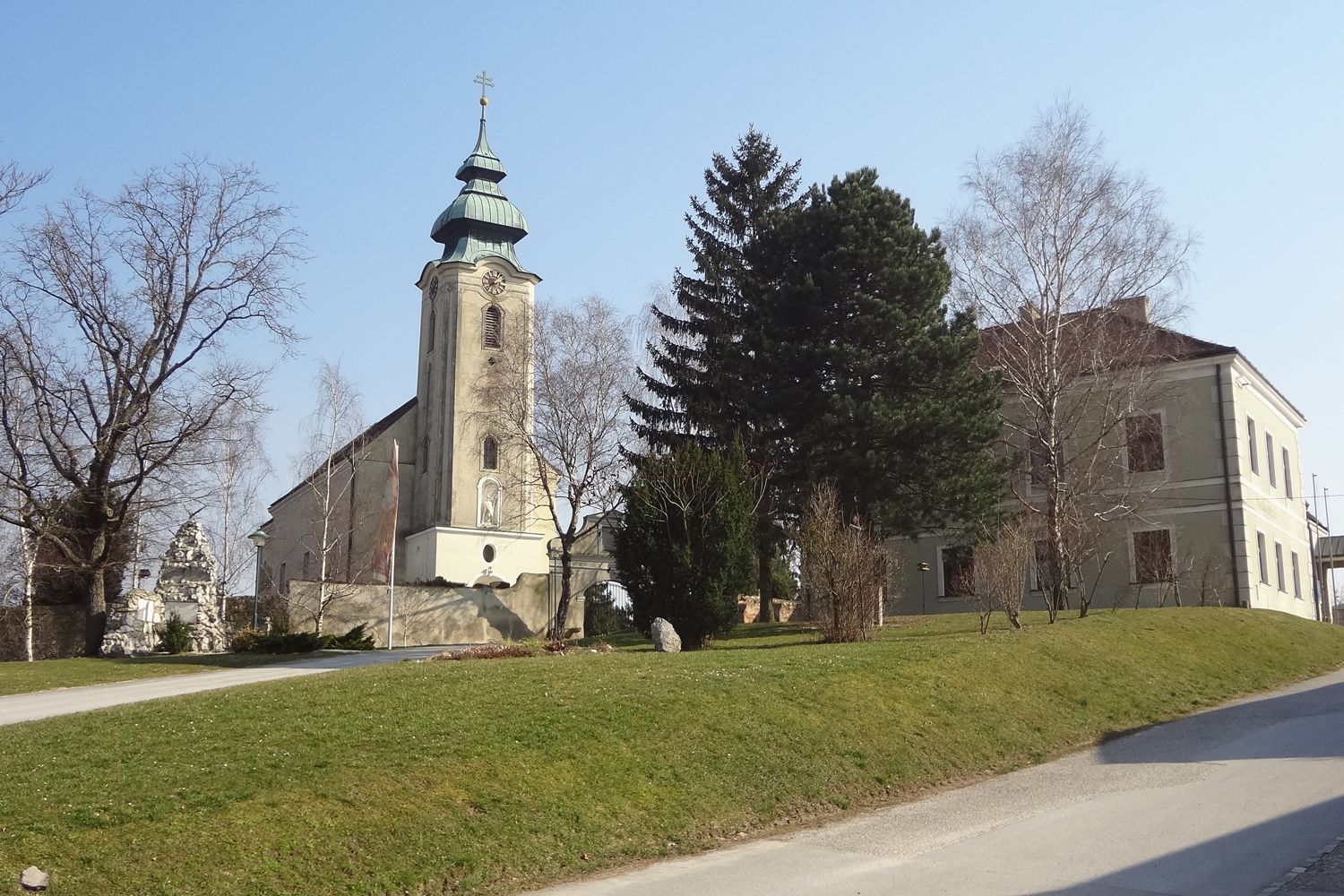 Kirche mit Turm und angrenzendem Gebäude in Gnadendorf, umgeben von Bäumen und Rasenfläche.
