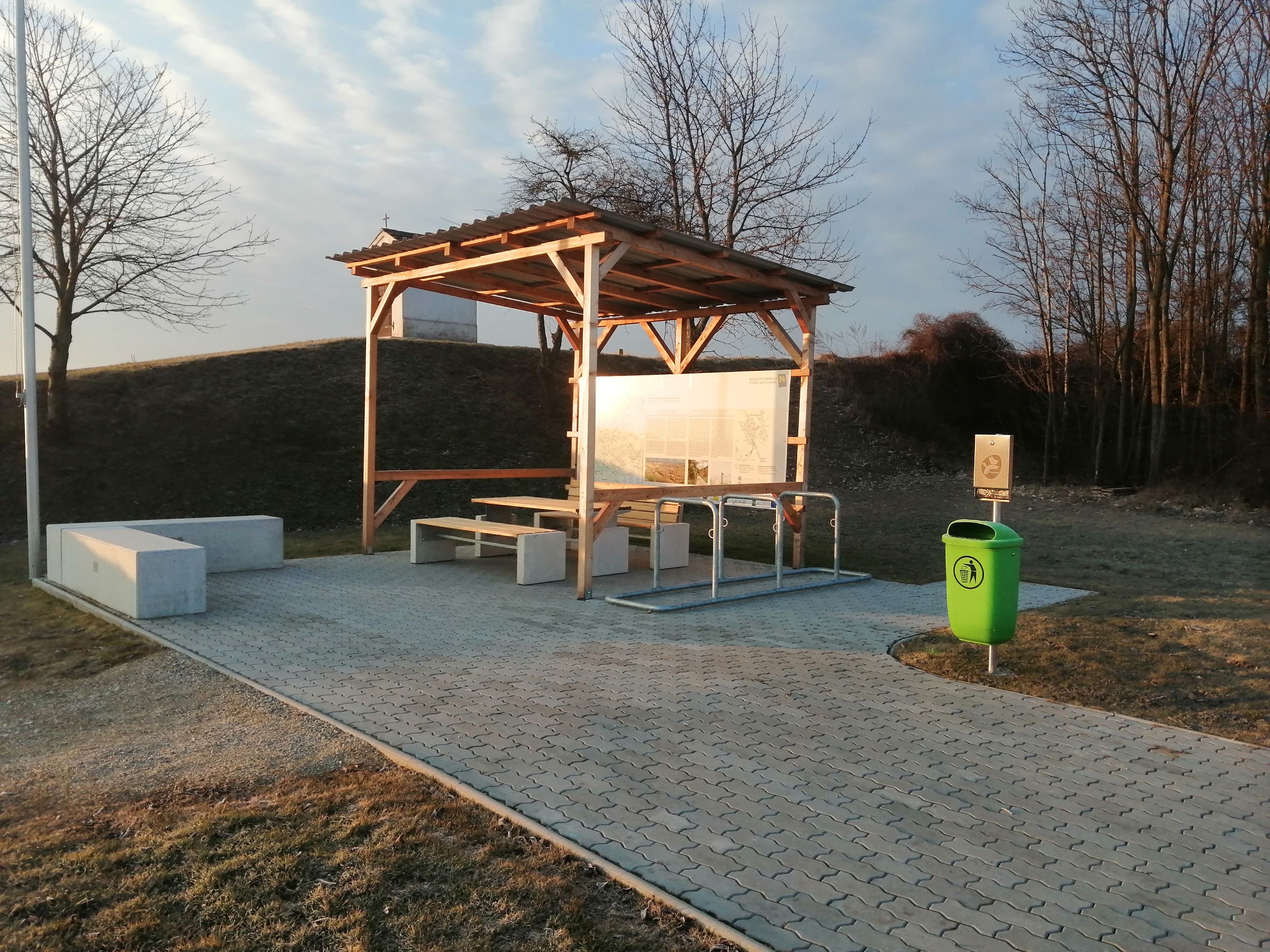 A rest area with a wooden roof, benches, and a green trash can on a paved surface, surrounded by bare trees and grass.
