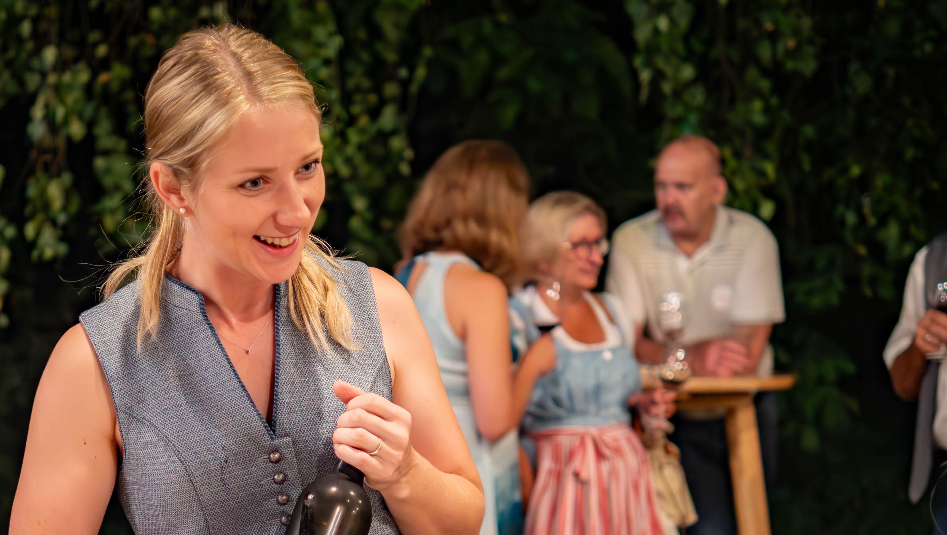 Woman with blond hair holding a bottle, in the background people with wine glasses.