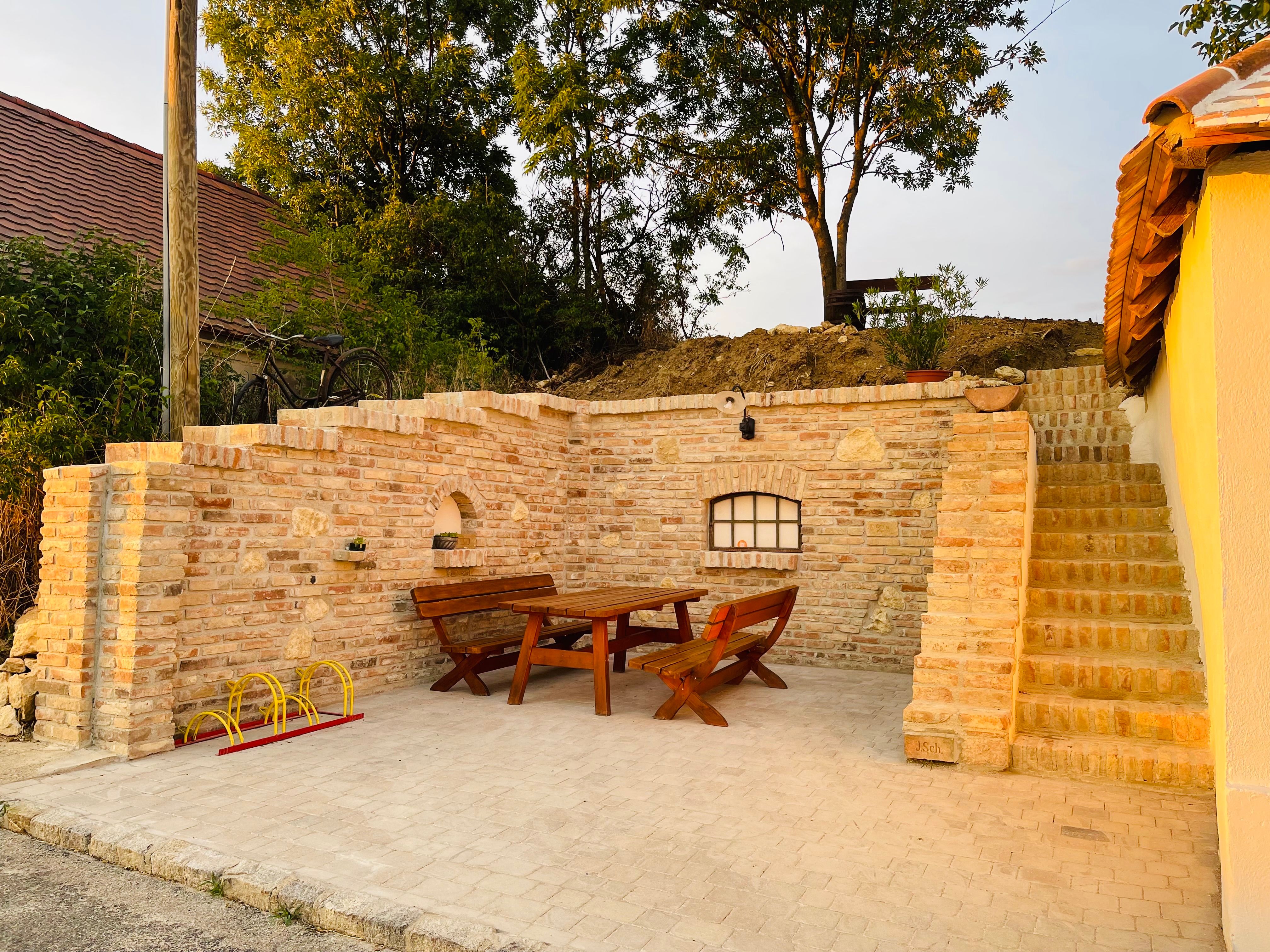Cozy outdoor area with wooden table and benches, surrounded by brick walls and stairs, in the evening light.