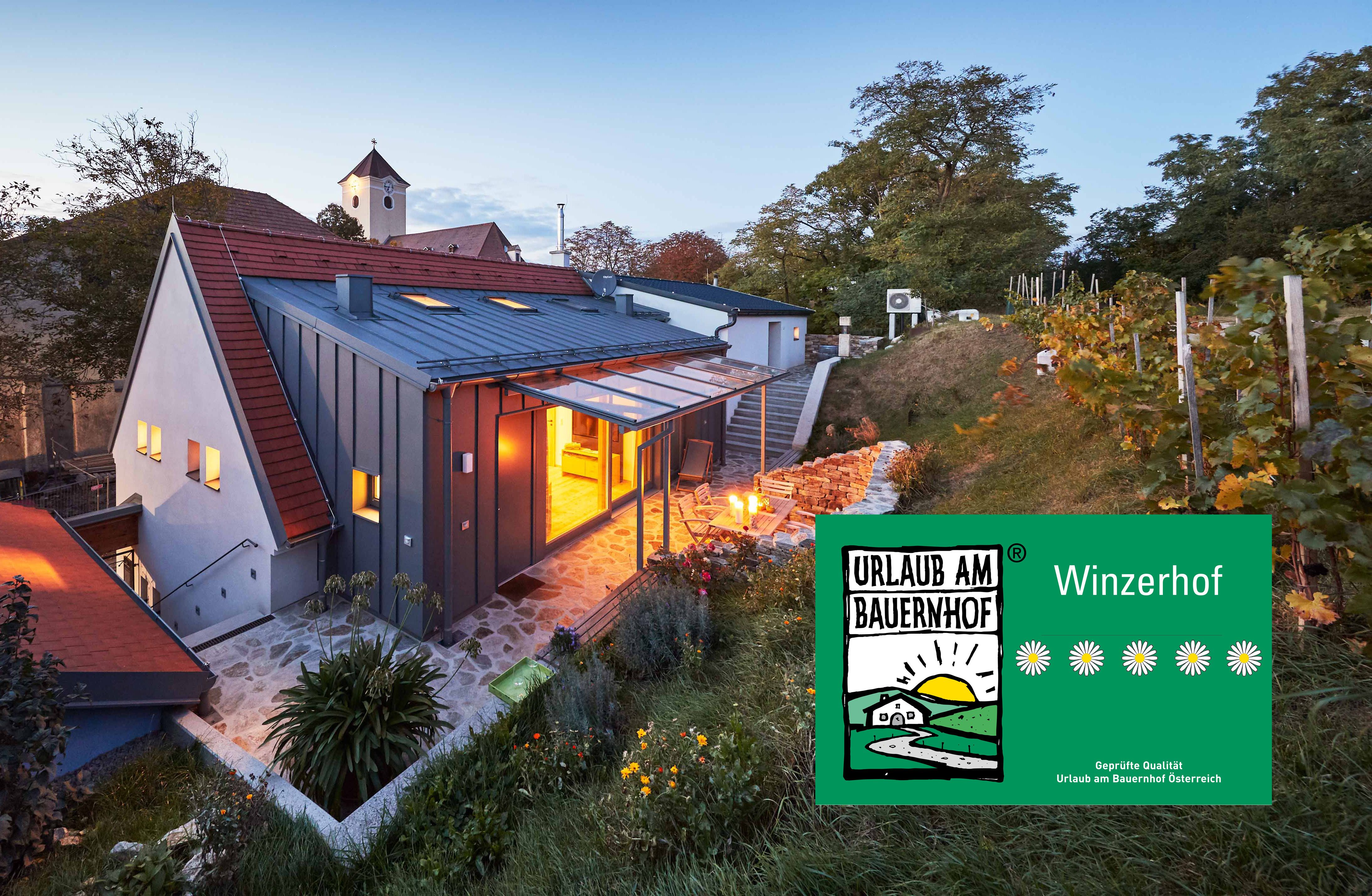 Ein modernes Kellerstöckl im Weinviertel bei Dämmerung, umgeben von Weinreben und einem Garten. Ein Schild zeigt 'Urlaub am Bauernhof'.
