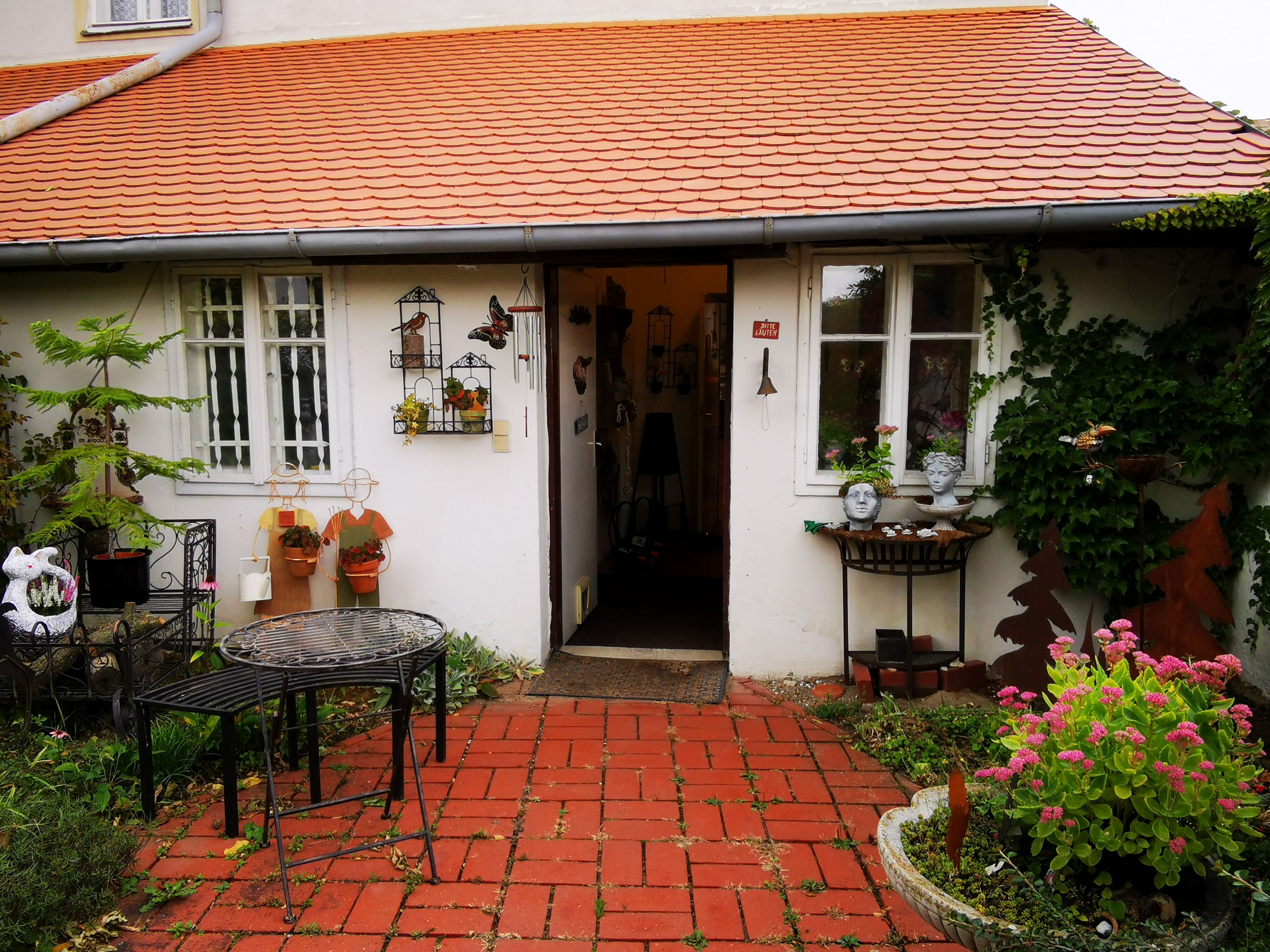 Eingang eines Hauses mit roten Dachziegeln, dekoriert mit Pflanzen und Keramikfiguren.
