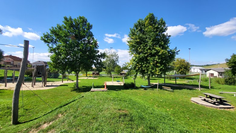 Ein Spielplatz mit Klettergerüst, Schaukeln und Picknicktisch auf einer grünen Wiese unter blauem Himmel.
