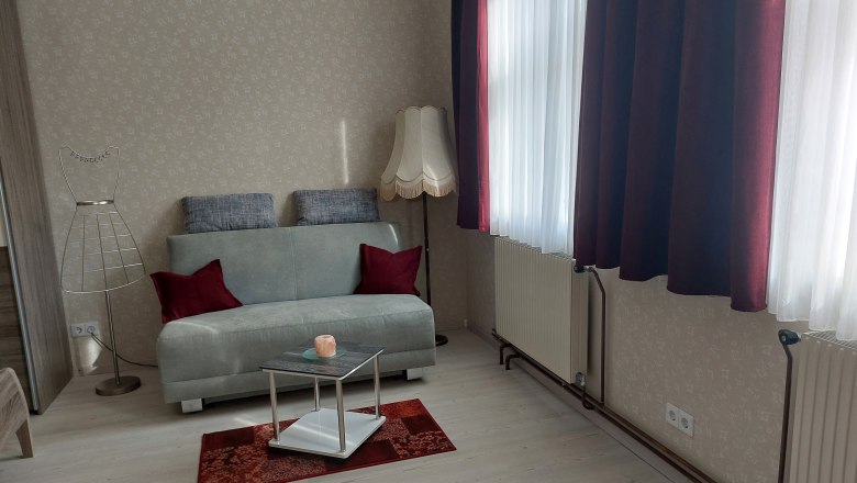 Wohnzimmer mit grauem Sofa, roten Kissen, Stehlampe und kleinem Tisch auf rotem Teppich.