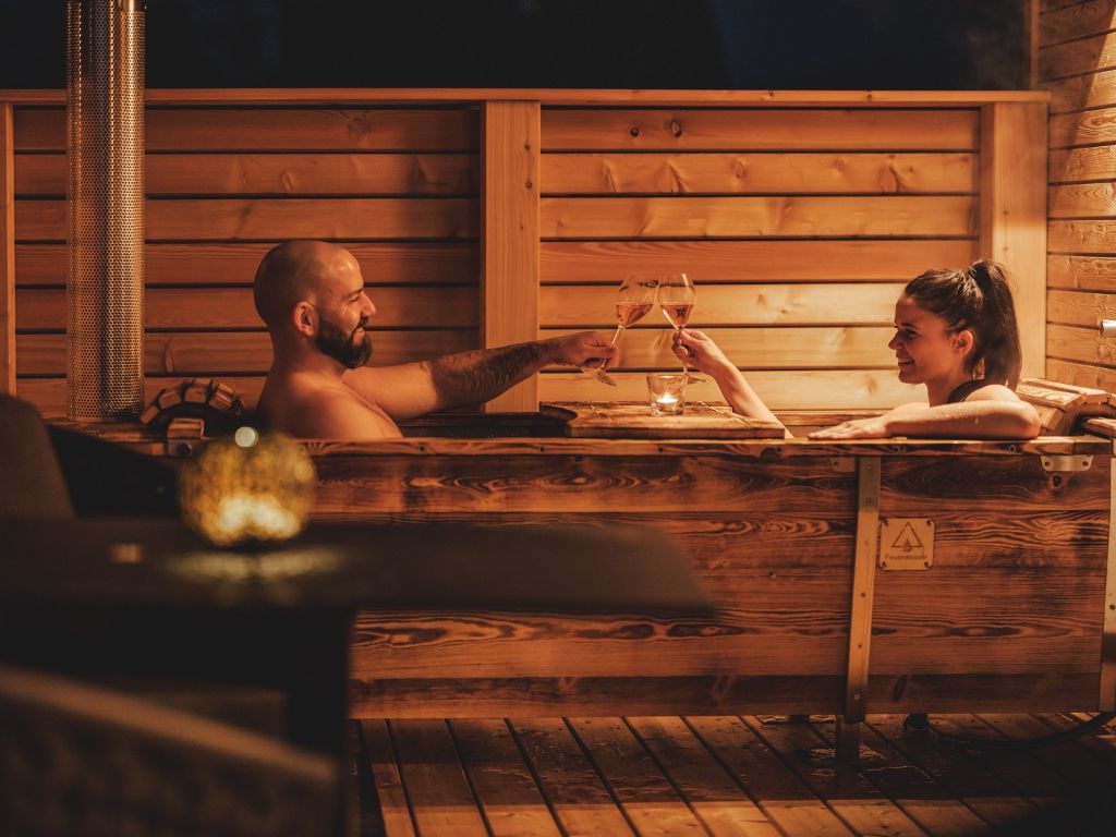Zwei Personen in einer Holzbadewanne im Freien, die mit Weingläsern anstoßen.