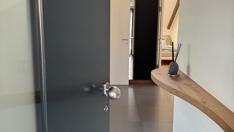 Moderner Eingangsbereich mit offener T&uuml;r, Fliesenboden und Holzregal an der Wand.