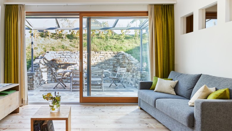 Innenansicht eines modernen Wohnzimmers mit grauem Sofa, Holzboden und großen Fenstern, die auf eine Terrasse mit Steinmauer und Gartenmöbeln führen.