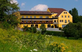 Gelbes Hotelgeb&auml;ude mit Blumenbalkonen, umgeben von gr&uuml;ner Landschaft.
