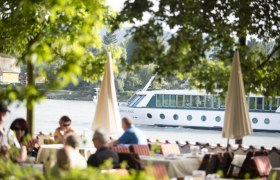 Ein Flusskreuzfahrtschiff fährt auf der Donau, im Vordergrund sitzen Menschen in einem Café unter Bäumen.