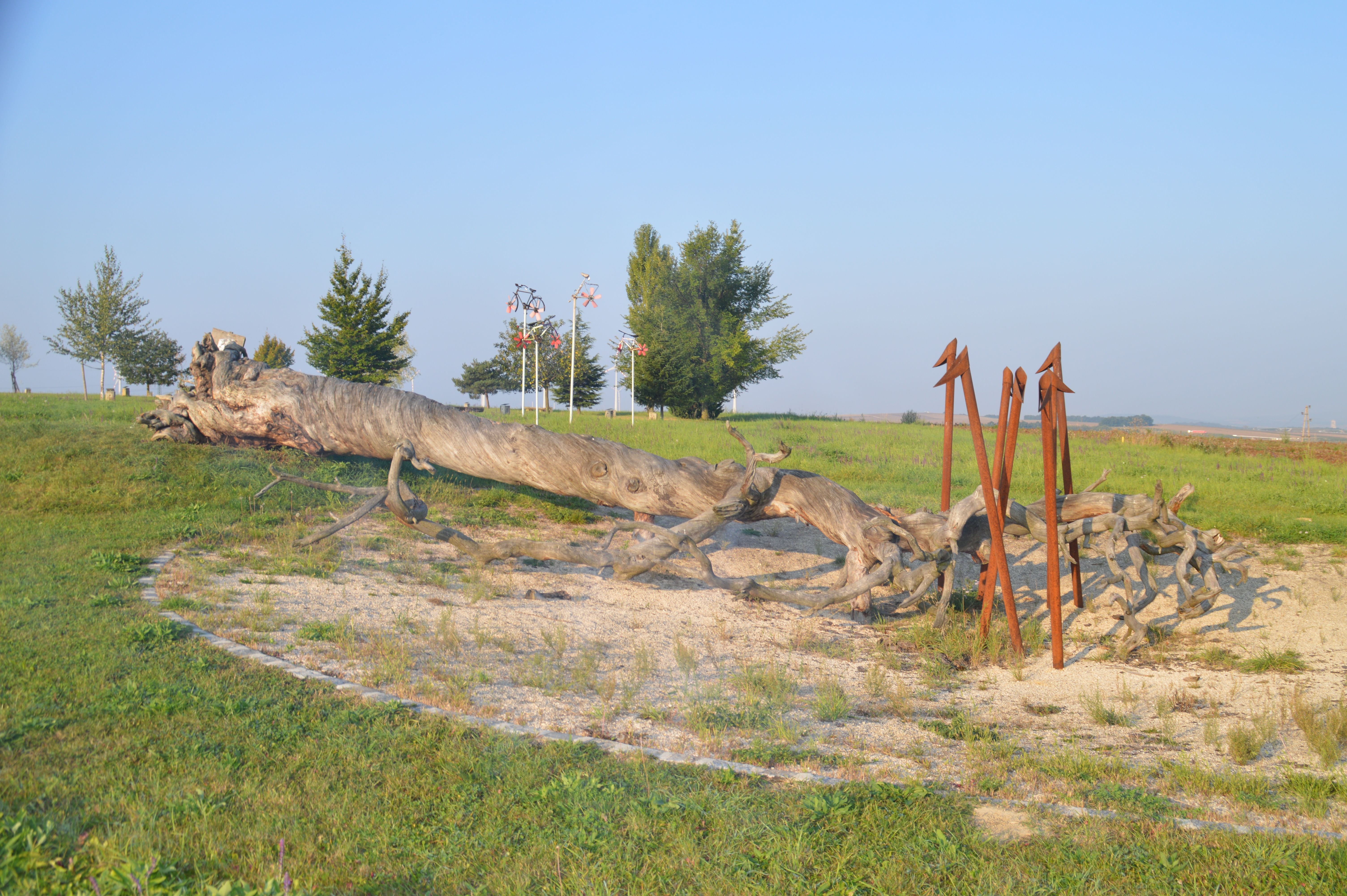 Ein großer, umgestürzter Baumstamm liegt auf einer Wiese, umgeben von Metallskulpturen und Bäumen im Hintergrund.