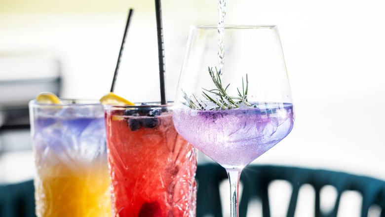 Drei bunte Cocktails auf einem Tisch, eine Hand gießt Flüssigkeit in ein Glas.