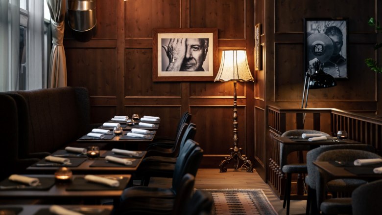 Gemütlicher Raum mit Holzwänden, gedeckten Tischen, Stehlampe und Schwarz-Weiß-Fotografien an der Wand.