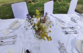 Gedeckter Tisch mit Blumenstrauß vor Weinberg und Liegestühlen.