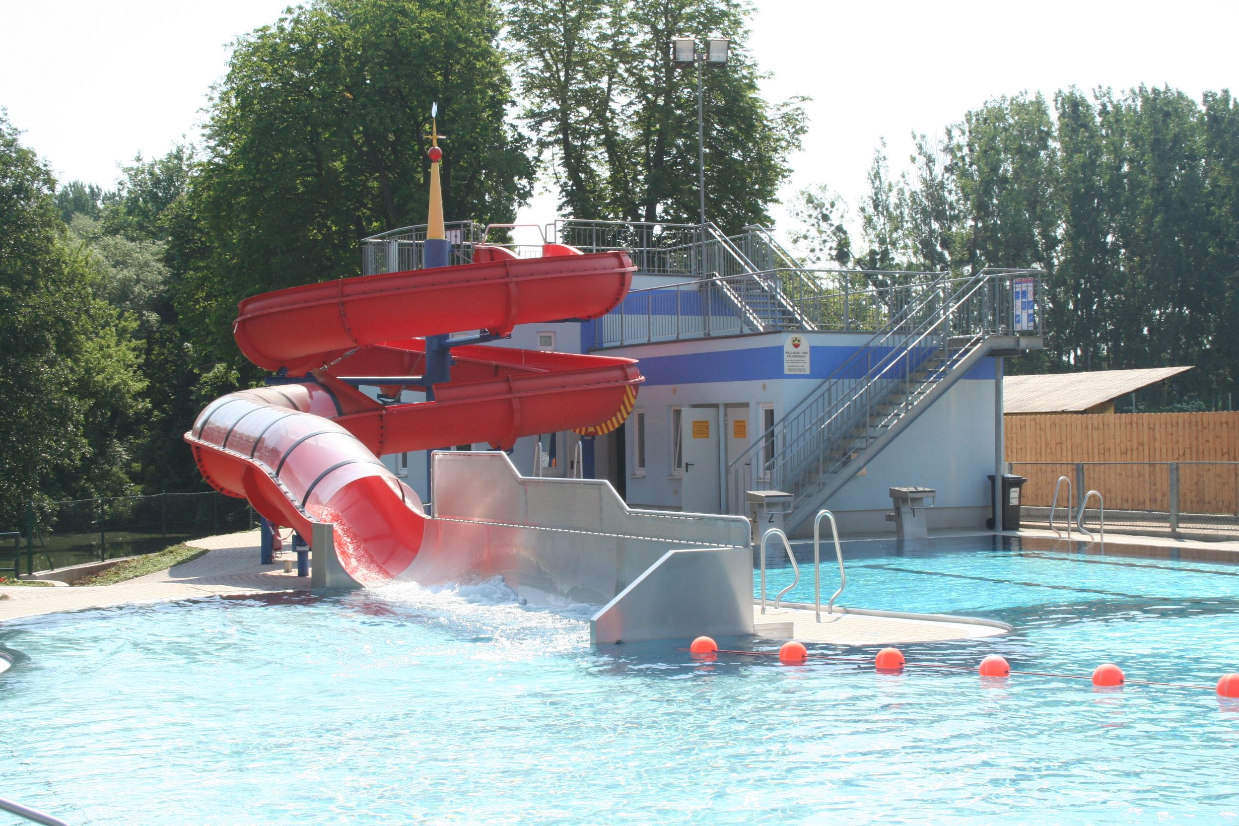 Rote Wasserrutsche in einem Freibad mit Pool und Bäumen im Hintergrund.