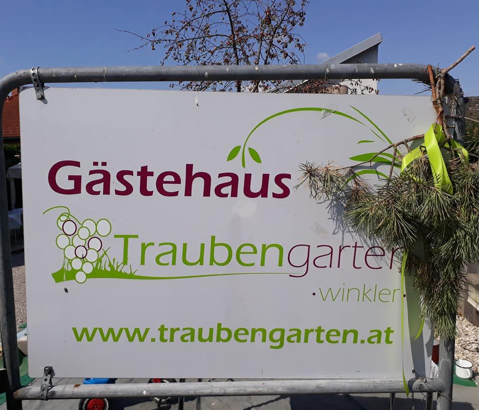 Schild mit der Aufschrift 'Gästehaus Traubengarten Winkler' und Website-Adresse, dekoriert mit Zweigen.