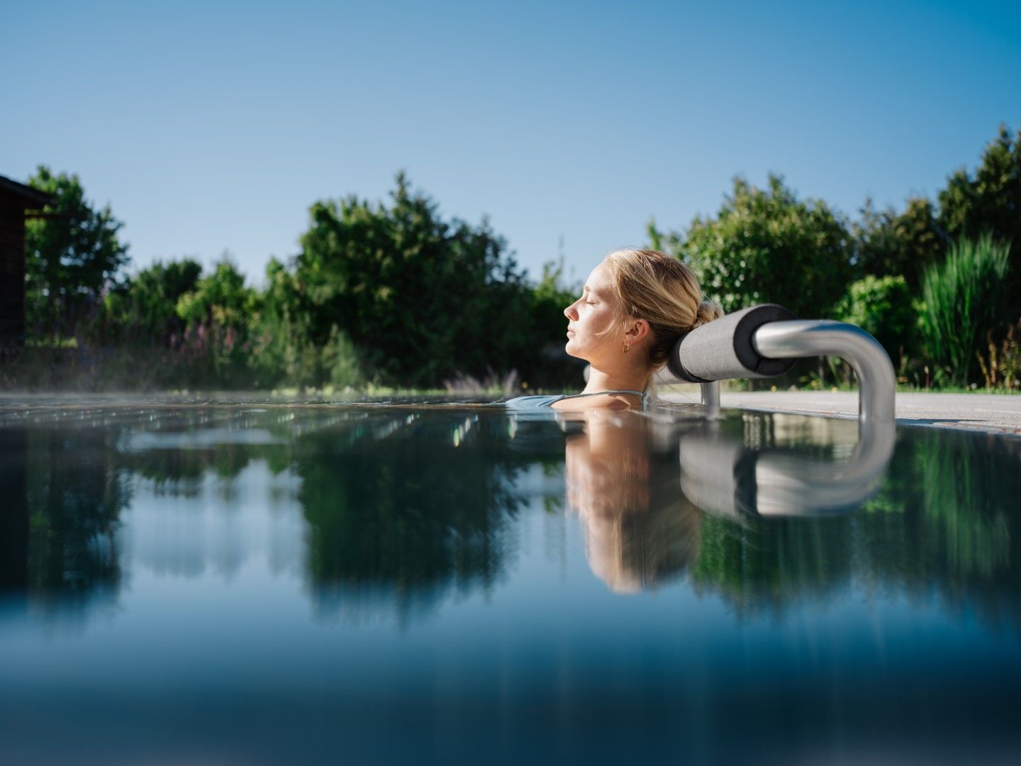 A woman relaxes in an outdoor pool, surrounded by green nature and clear skies.
