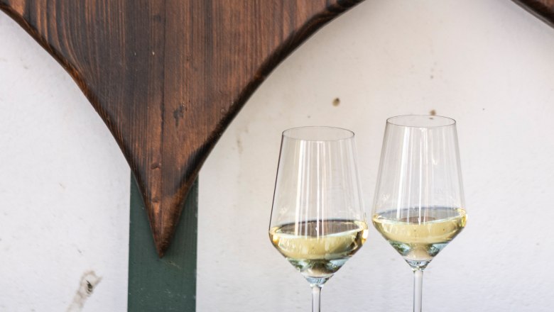 Two wine glasses with white wine on a wooden table in front of a wooden wall with heart decoration.