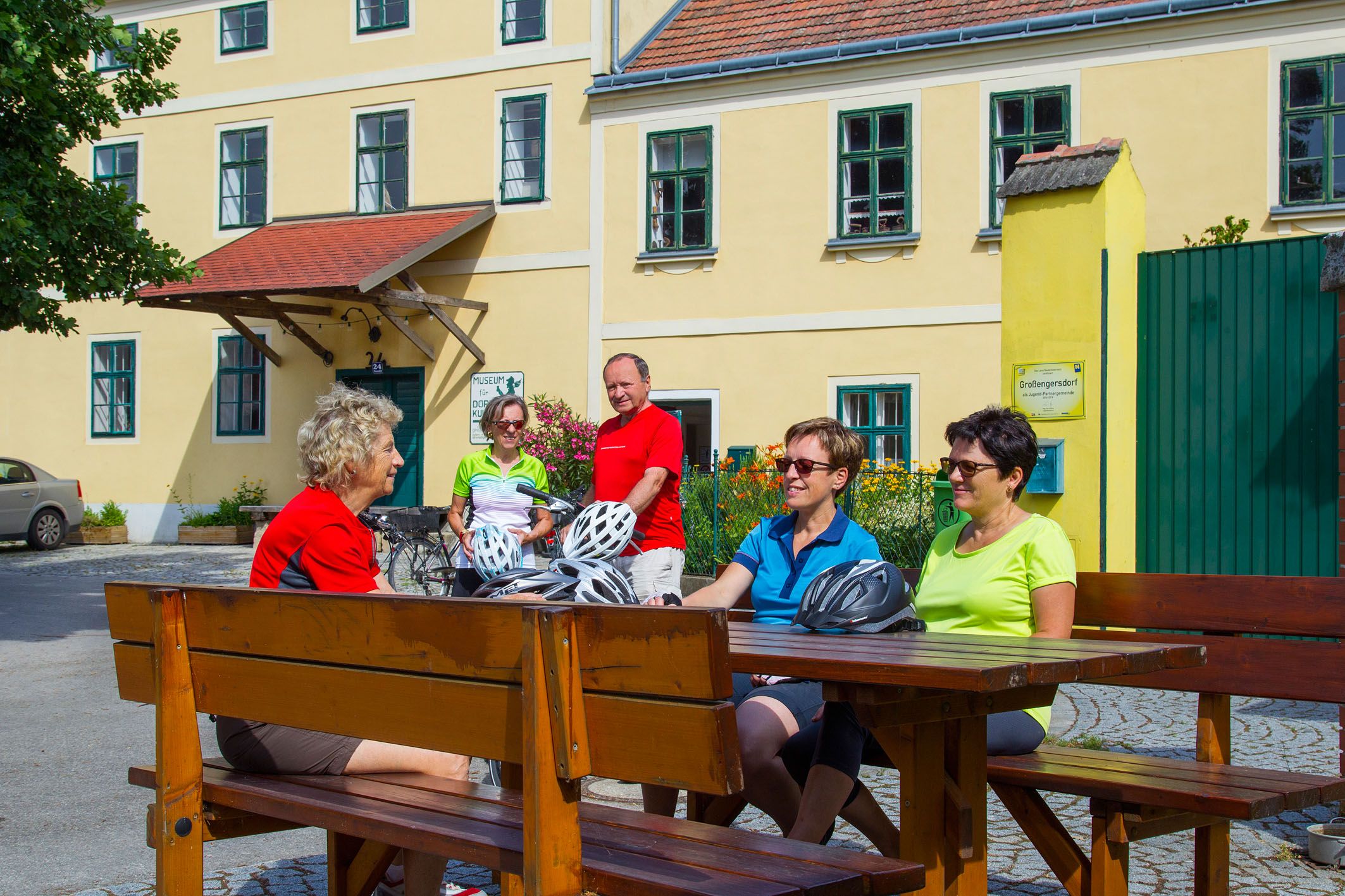 Gruppe von Radfahrern sitzt an einem Holztisch vor einem gelben Gebäude.