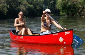 Zwei Frauen paddeln in einem roten Kanu auf einem Fluss, umgeben von grüner Natur.