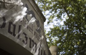 Nahaufnahme eines alten Geb&auml;udes mit Schriftzug und Wappen, umgeben von B&auml;umen.