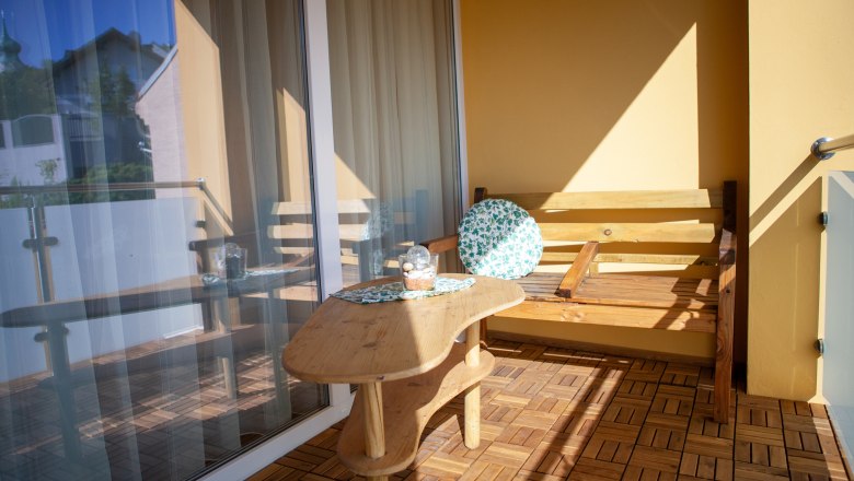 Ein Balkon mit Holzboden, einem Holztisch und einer Holzbank mit einem runden Kissen. Im Hintergrund ist ein Glasgeländer und ein Gebäude zu sehen.