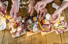 Mehrere Hände greifen nach Wurst- und Käseplatten auf einem Holztisch.