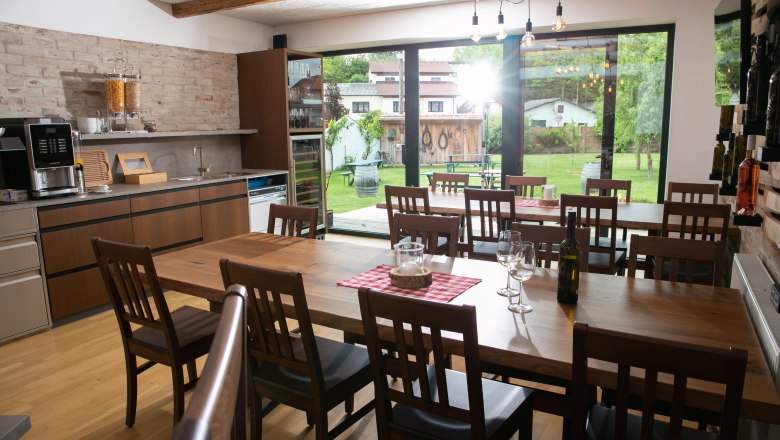 Innenansicht eines modernen Gastraums mit Holztischen und St&uuml;hlen, gro&szlig;en Fenstern und Blick auf einen Garten.