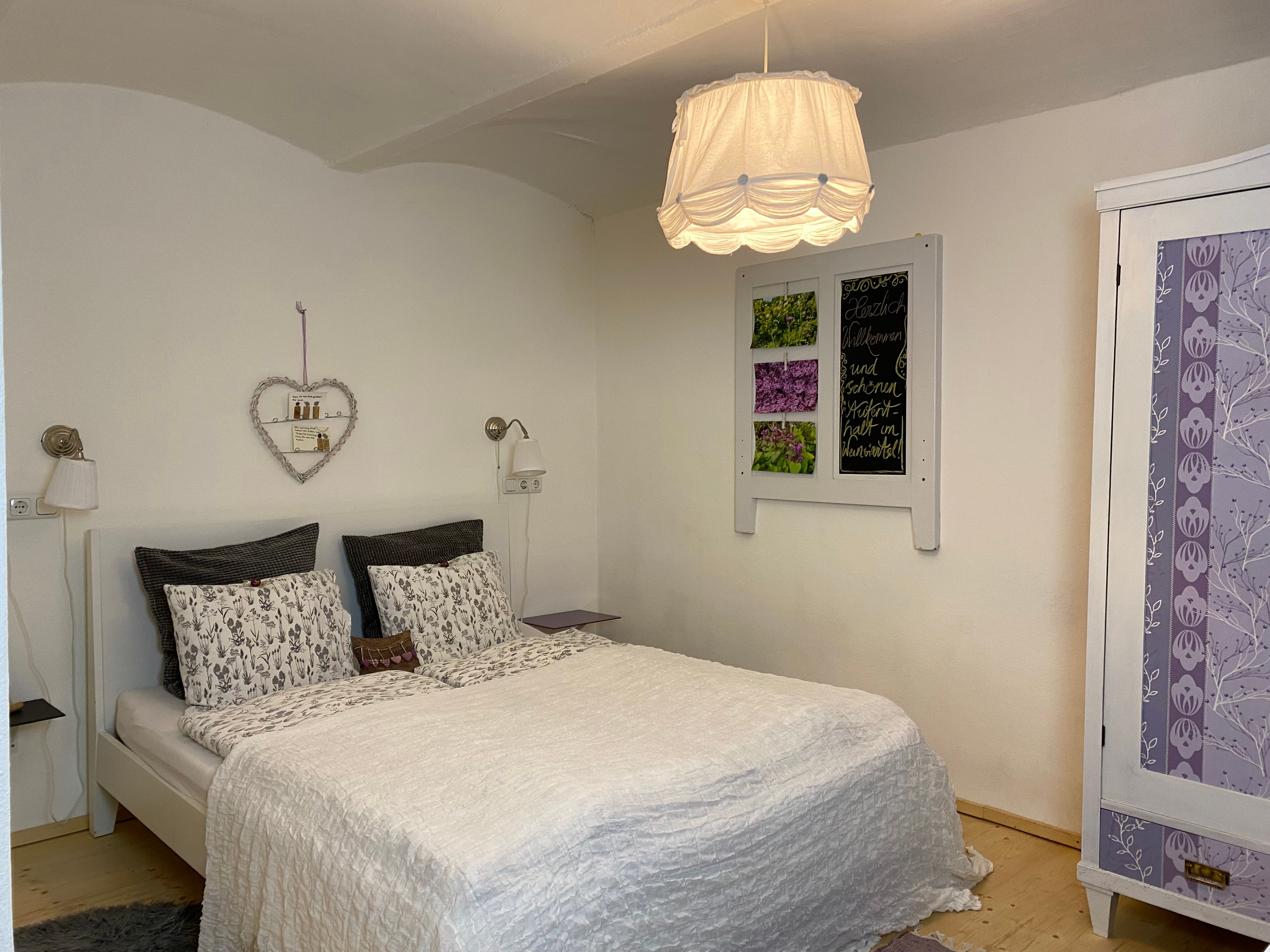 Gemütliches Doppelzimmer mit weißem Bett, dekorativem Herzregal und lila Schrank.