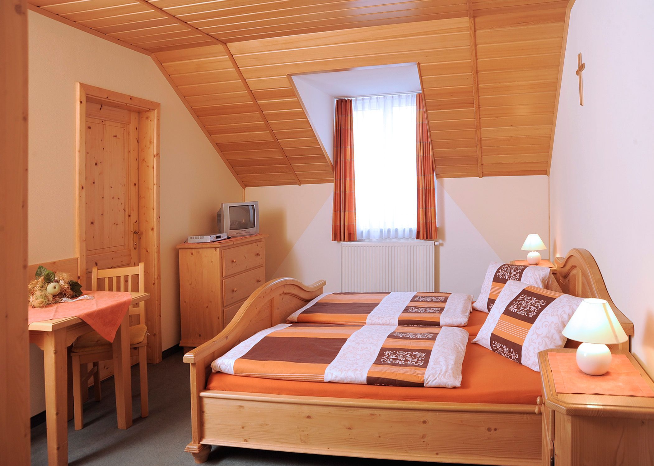 Gemütliches Gästezimmer mit Holzmöbeln, Doppelbett, kleinem Tisch, Fernseher und Dachschräge.