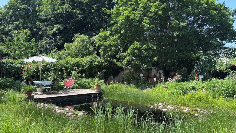 Ein idyllischer Garten mit Teich, Holzterrasse, Tisch und Stühlen unter einem Sonnenschirm, umgeben von üppiger Vegetation und Bäumen.