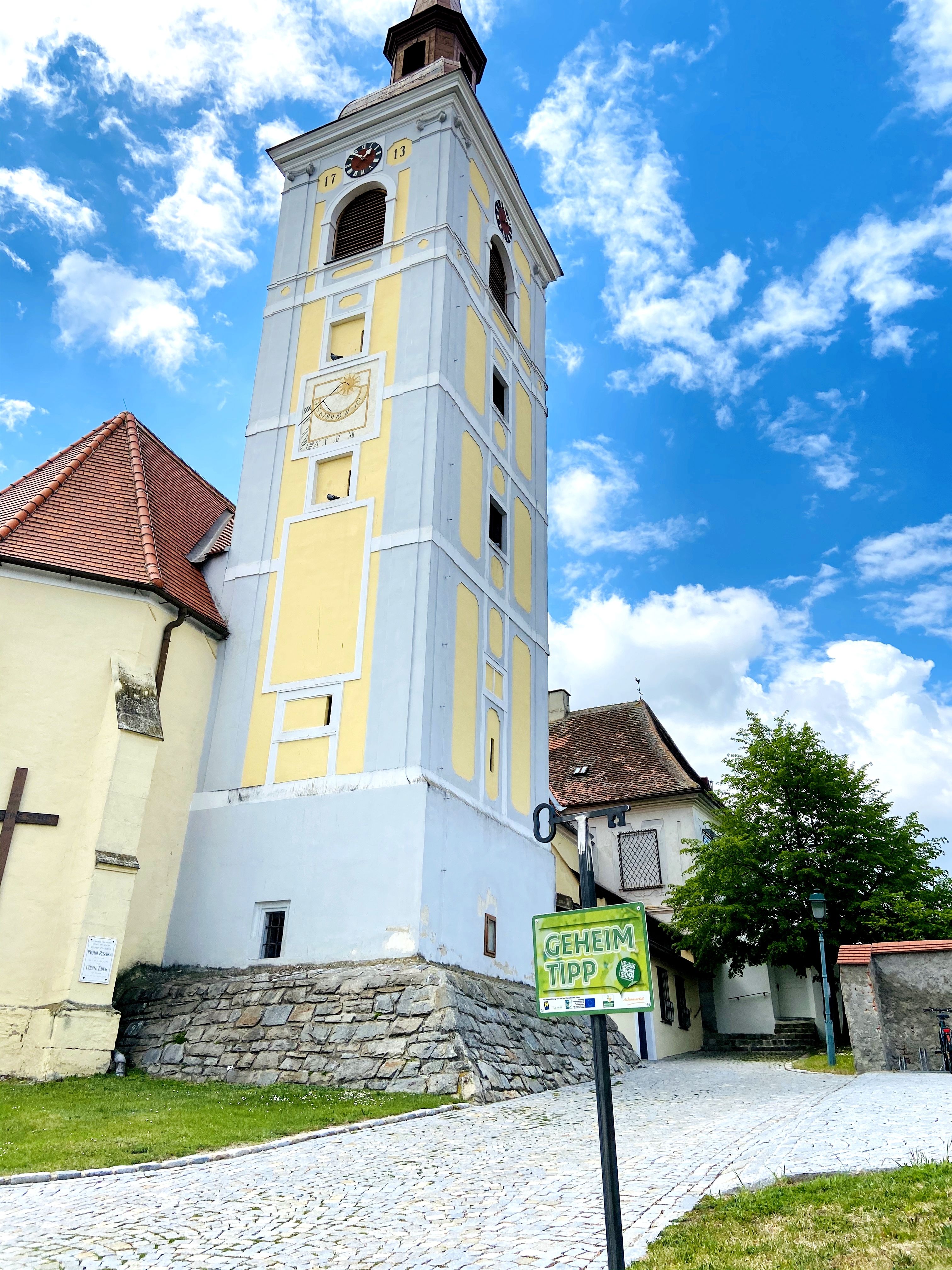 Schiefer Turm in Waitzendorf mit gelber Fassade und Uhr, umgeben von blauen Himmel und Wolken.