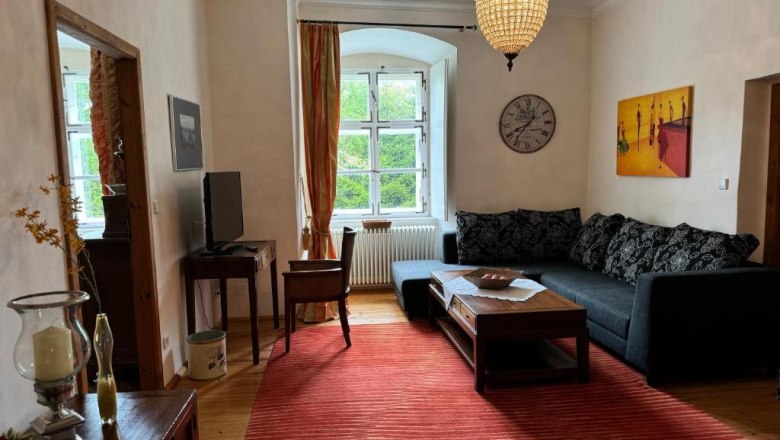 Gemütliches Wohnzimmer mit Sofa, rotem Teppich, Kronleuchter und großem Fenster.