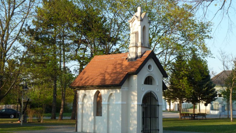 Eine kleine weiße Kapelle mit rotem Dach in einem Park, umgeben von Bäumen.