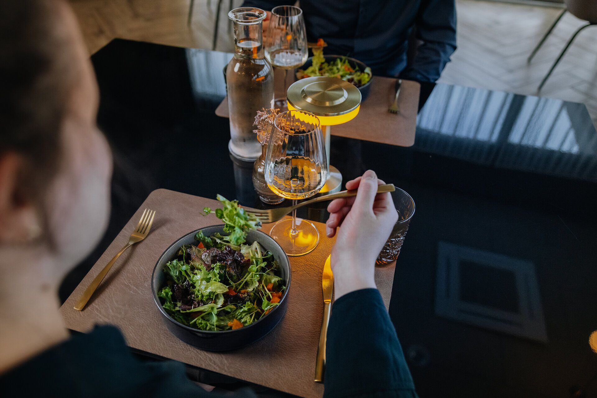 Eine Frau sitzt in einem Restaurant an einem Tisch und isst einen Salat. Vor dem Teller stehen ein Wasserkrug, ein Glas Wasser sowie ein Glas Weißwein, der Blick fällt über ihre Schulter.