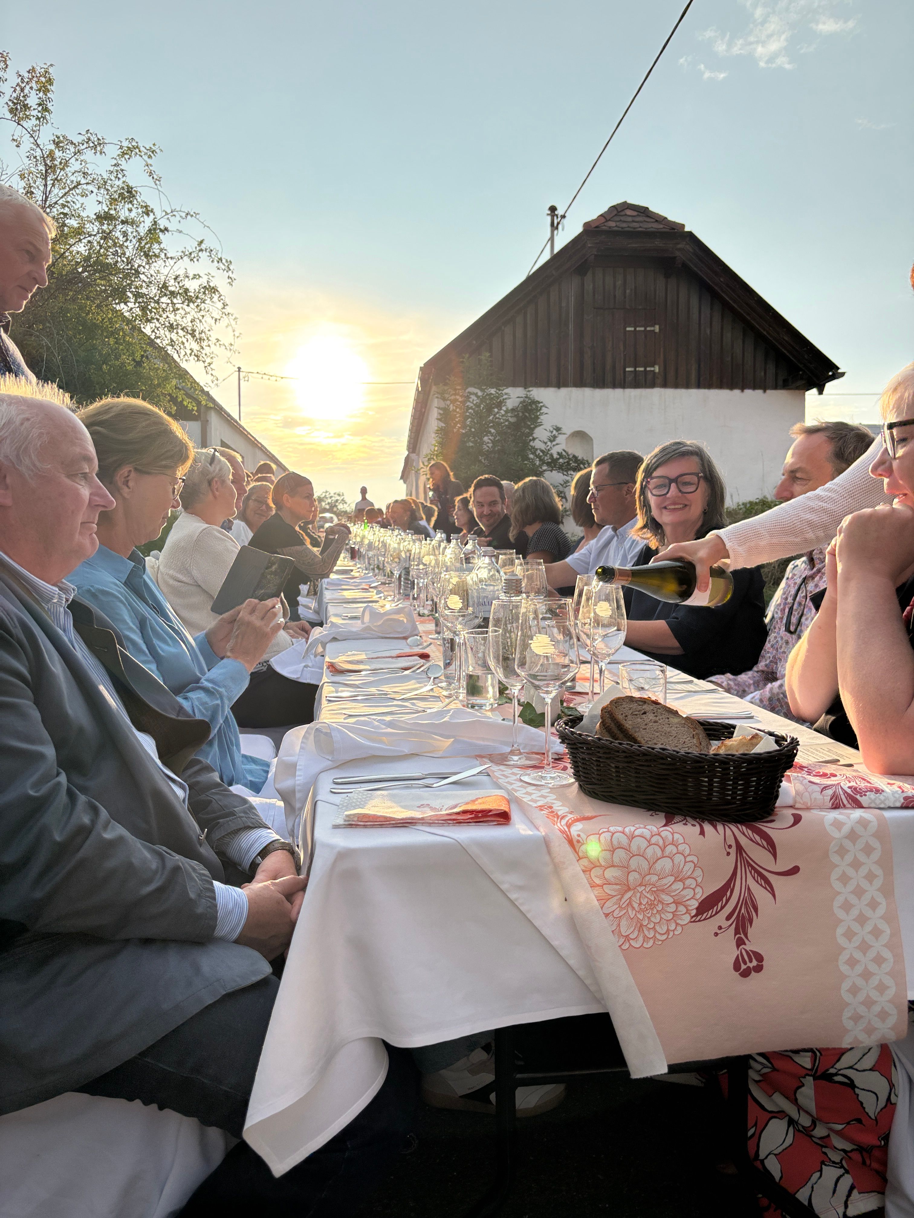 Menschen sitzen an einer langen Tafel im Freien bei Sonnenuntergang.