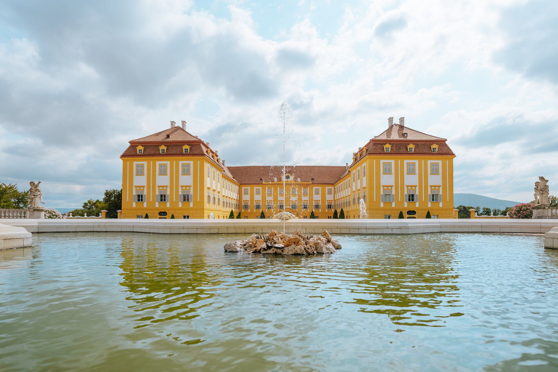 Die majestätische Fassade des Schlosses Hof strahlt in warmen Gelbtönen und spiegelt sich im ruhigen Wasser des Brunnens wider. Umgeben von üppigen Gärten und Terrassen lädt dieser Ort dazu ein, die Schönheit der Natur und die Pracht der Architektur zu genießen.