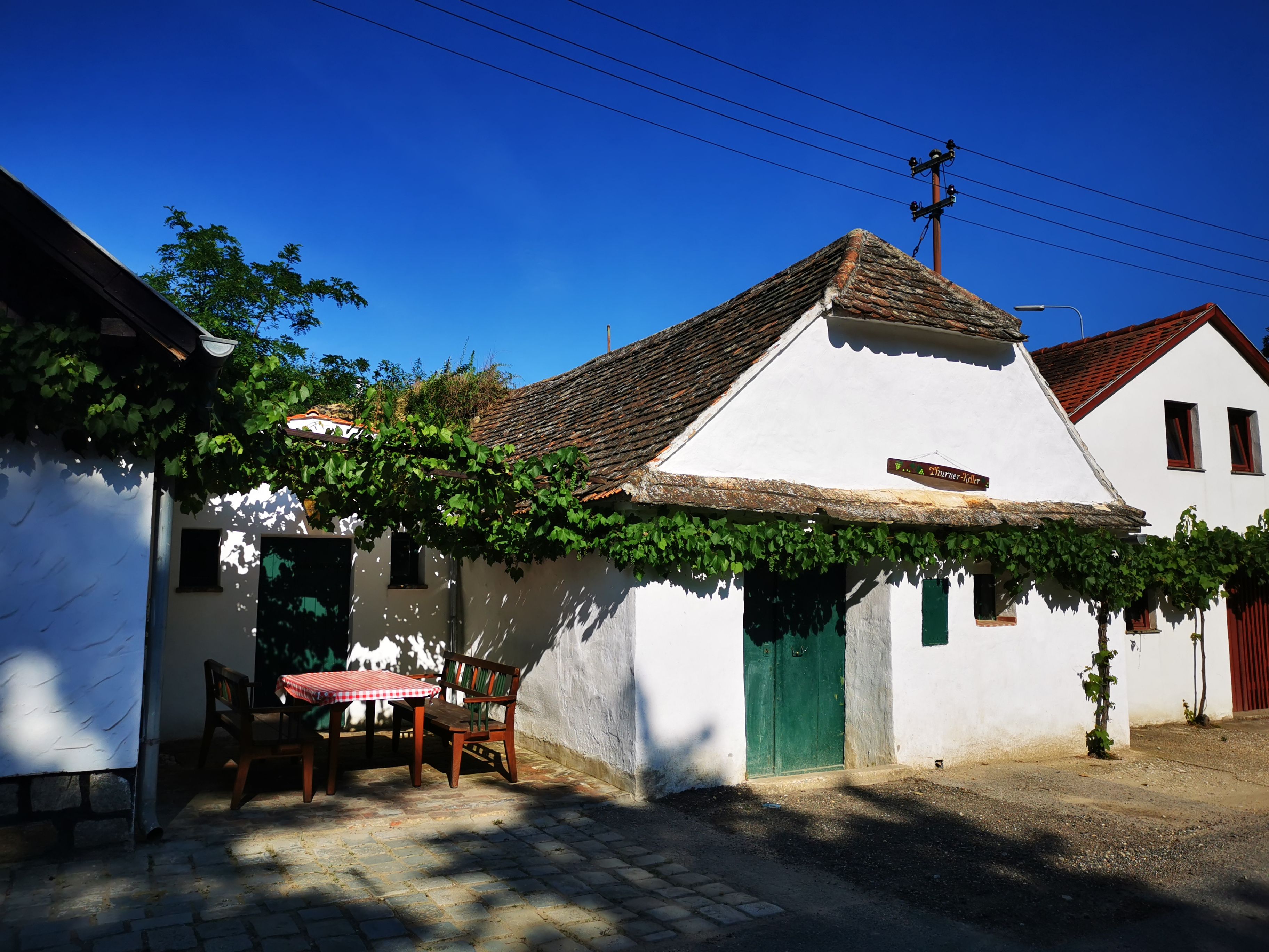 White building in a wine cellar lane with vines and outdoor table.