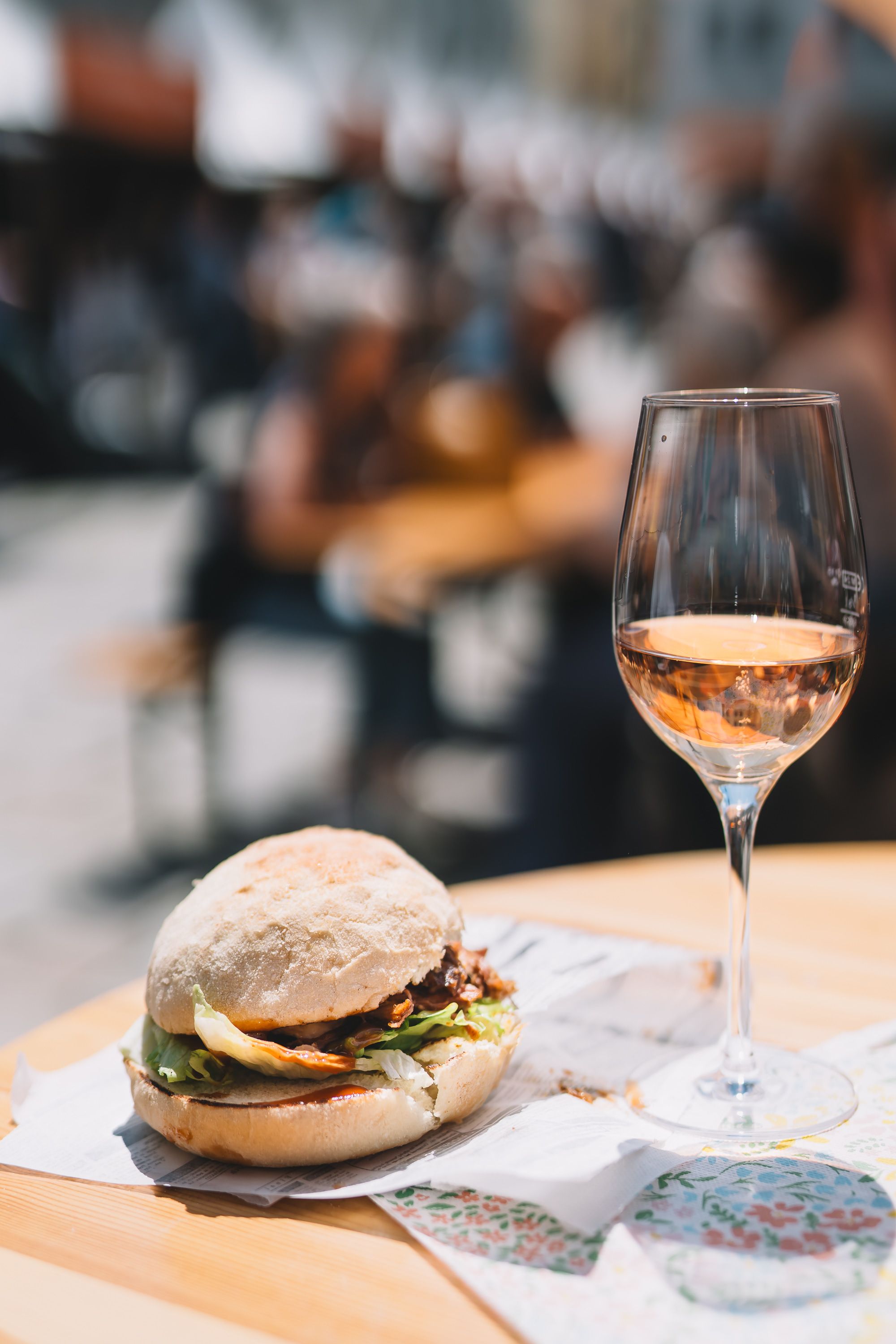 Ein Glas Rosé und ein Weinviertler Burger stehen auf einem Stehtisch, im Hintergrund genießen Personen das Fest.