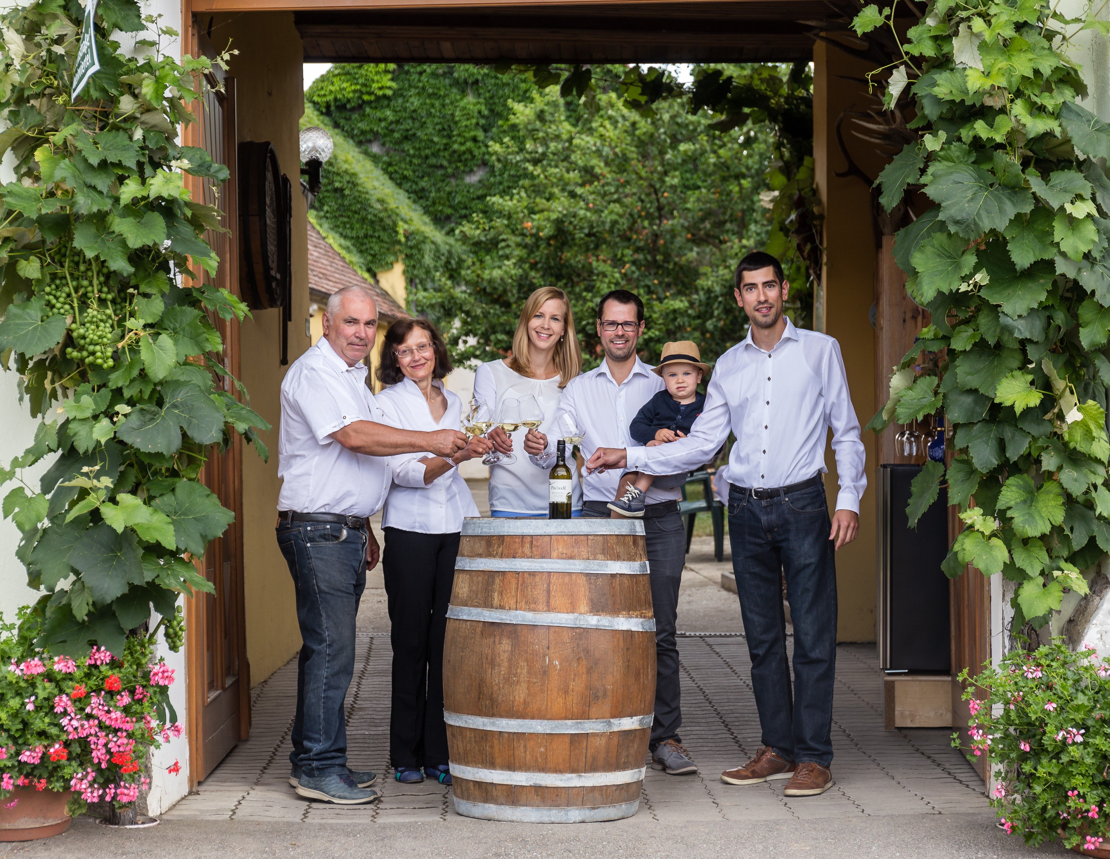 Group of people standing around a wine barrel, holding wine glasses and smiling at the camera. Surrounded by vines and flowers.